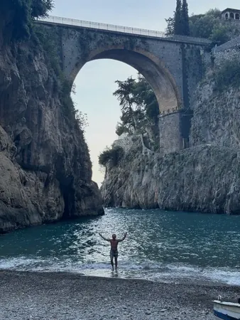 A swimmer standing arms wide on a narrow beach under a stone arch on the coast